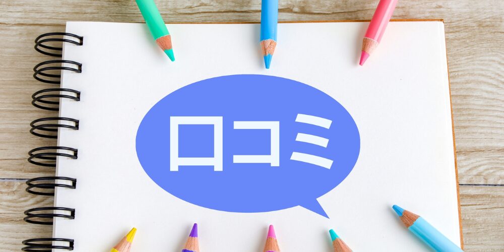 白いリングノートの上に青い吹き出しで「口コミ」と大きく書かれた文字が乗り、その周囲に色鉛筆が並ぶ、口コミ紹介を示すイメージ写真。