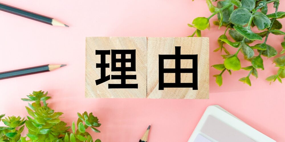 ピンクの背景に置かれた木製ブロックに「理由」と書かれた文字。観葉植物と文具が周囲に配置されている