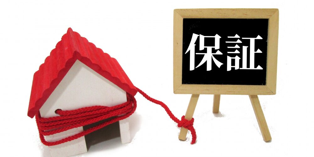 赤い屋根の模型住宅が赤い紐で結ばれ、「保証」と書かれた黒板が横に置かれている写真。保証の安心感を表現