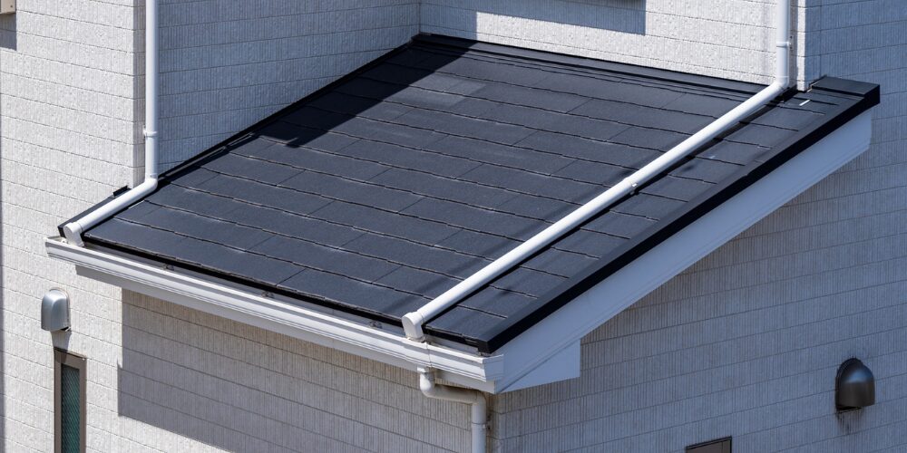 白い外壁に取り付けられた黒いタイル葺きの屋根部分をクローズアップ。雨樋や縁の金具が見え、強い日差しを浴びたシャープな屋根のディテールを写したイメージ