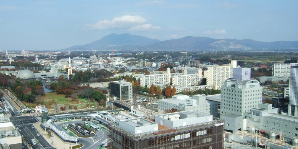 晴天の下、広がる市街地のビル群と駅周辺、高層マンション郡の背後に緑の山並みが連なるパノラマ風景写真。