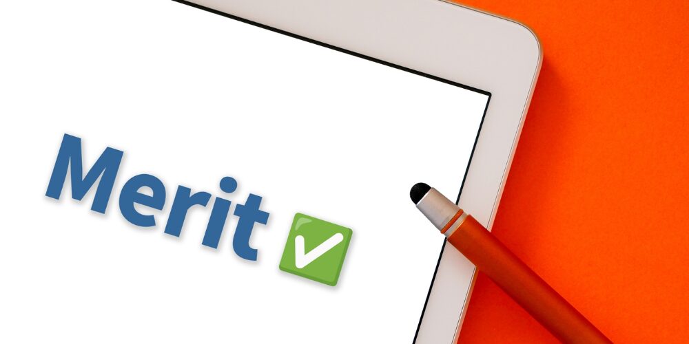 オレンジの背景にタブレット端末の白い画面上へ青い立体文字で「Merit✅」と表示され、横にオレンジのタッチペンが置かれた写真。