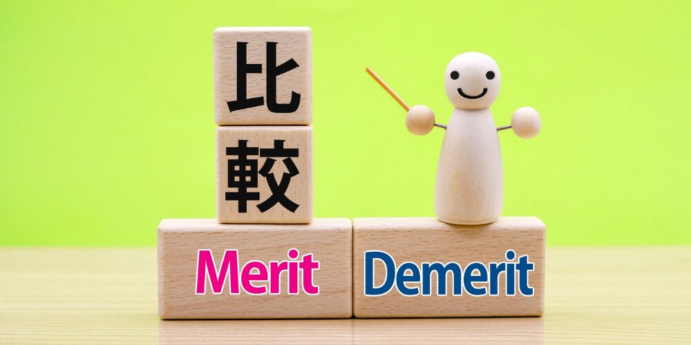 緑の背景に木製ブロックが積まれ、上段に黒文字で『比較』、下段左右に『Merit』（左、ピンク）と『Demerit』（右、青）、右側ににこやかな木製人形が指揮棒を持って立っているイメージ