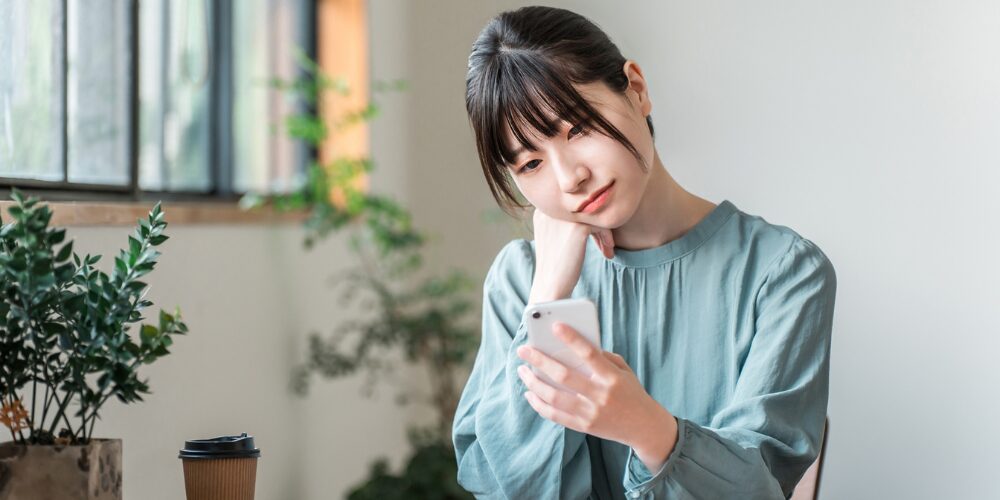 屋内のテーブル席で、青緑色のブラウスを着た若い女性がスマートフォンを手に取り、顎に手を当てながら画面を真剣に見つめているイメージ