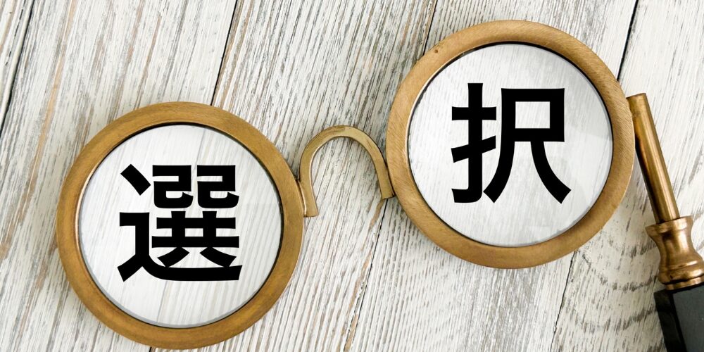 木製の丸眼鏡が開かれた状態でテーブルに置かれ、レンズ部分にそれぞれ「選」「択」の文字が拡大して見える画像