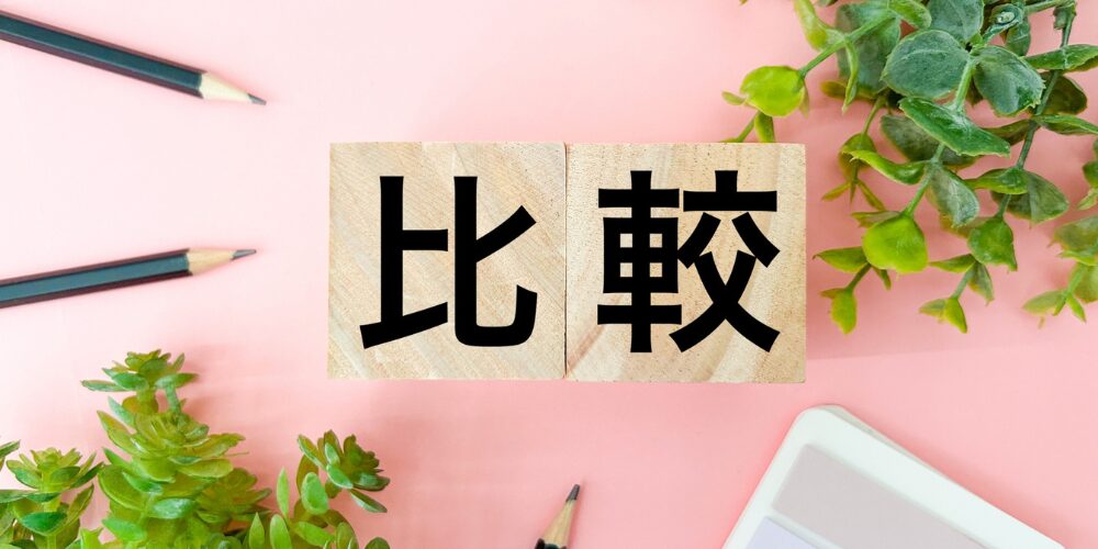 ピンクの背景に木製ブロックが横置きされ、黒文字で『比較』と書かれており、周囲に観葉植物と鉛筆が配置されたイメージ