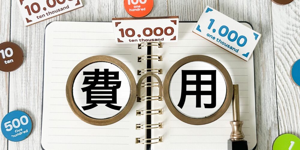 木目テーブルの上に開いたリングノートがあり、その上に２つの虫眼鏡が並べられている。左のレンズ内に黒文字で『費』、右に『用』と書かれている。周囲に紙製の小さな硬貨や札が散らばっているイメージ