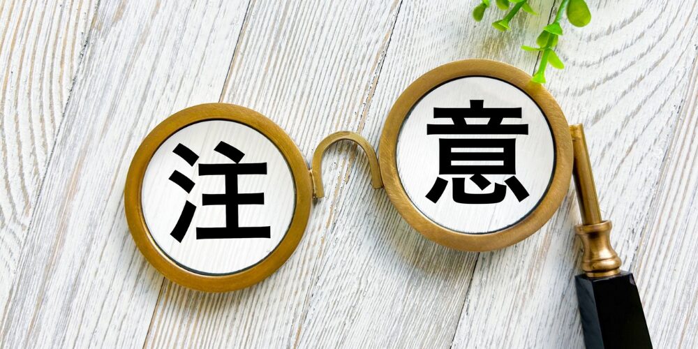 白木のテーブル板の上にアンティーク調の虫眼鏡が置かれ、そのレンズ内に黒文字で『注』（左）、『意』（右）と書かれている。背景に緑の観葉植物が見えるイメージ