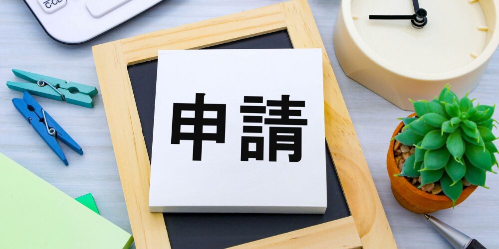 机の上に置かれた小さなホワイトボードのメモパッドの上に「申請」と書かれた一枚の紙が載っており、横に多肉植物や時計が写っている写真