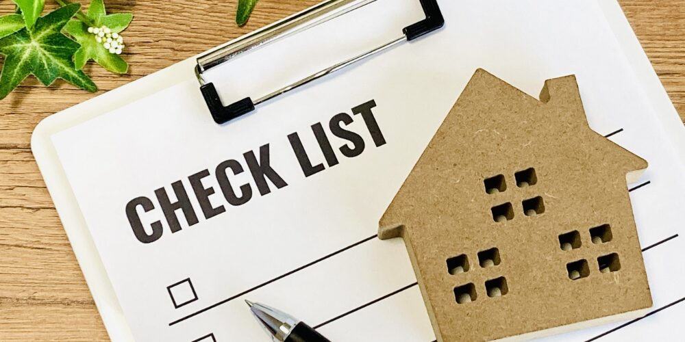 木目のテーブルに白いクリップボードが置かれ、『CHECK LIST』とタイトルが黒文字で印刷され、その上にペンと小さな木製の家型オブジェが並ぶイメージ