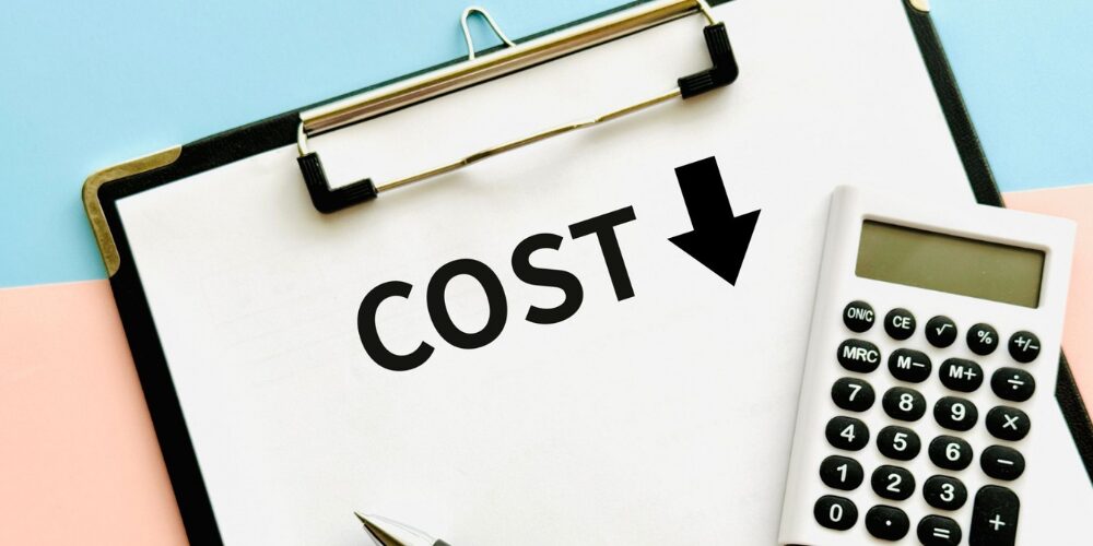 パステルブルーとピンクの背景の上にクリップボードが置かれ、白い用紙に『COST』と下向き矢印が黒文字で書かれ、右側に電卓とペンが並んでいるイメージ