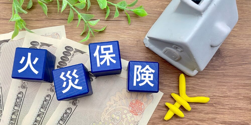 ALT　木目のテーブル上に千円札や一万円札が広げられ、その上に青い木製キューブが並び、白文字で『火』『災』『保』『険』と一つずつ刻まれている。隣に小さな家型オブジェと黄色い矢印オブジェが置かれているイメージ